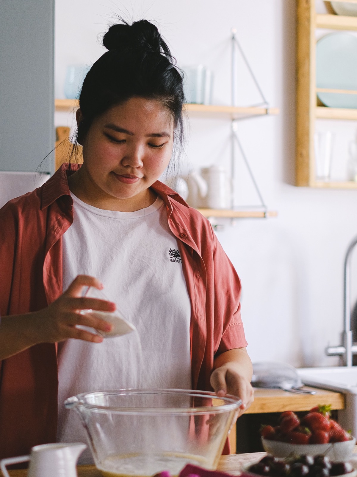 A woman in a home kitchen pouring an ingredient into a mixing bowl, with eggs and fresh fruit on the counter.